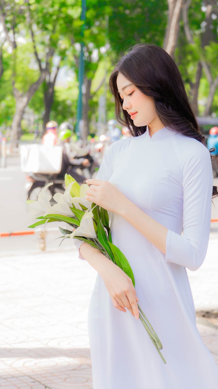 A serene portrait of an Asian woman in a white ao dai holding flowers on a sunny day outdoors.