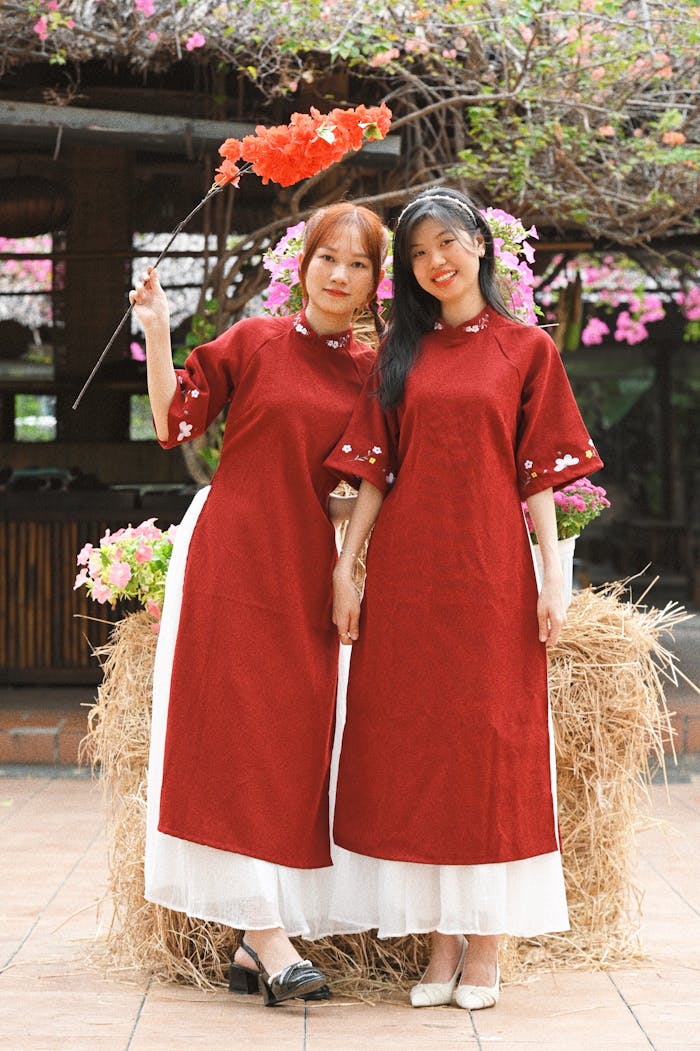 Two women in traditional red Áo dài dresses pose outdoors in Thành phố Hồ Chí Minh, surrounded by flowers.