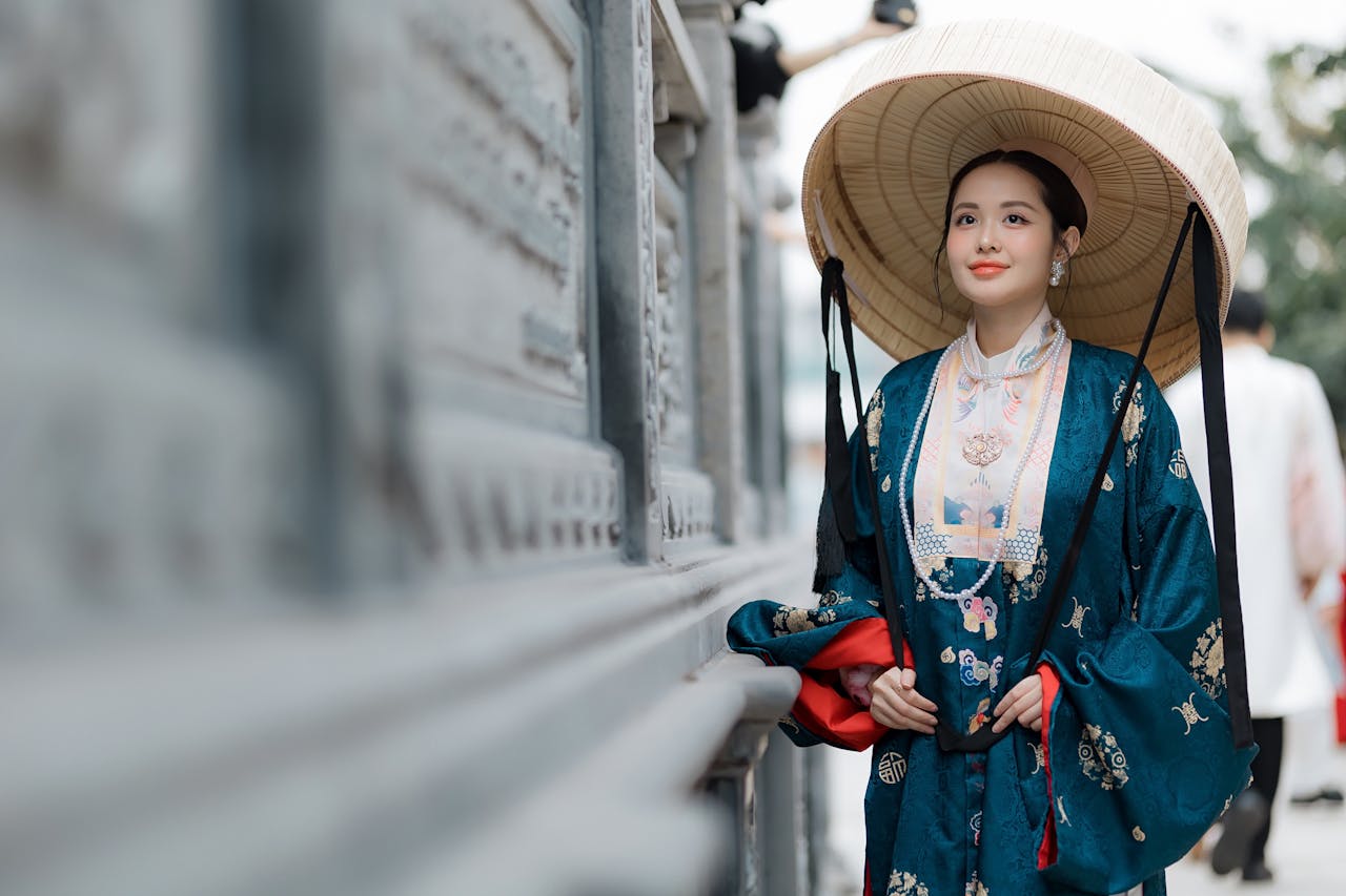 Elegant woman in traditional Asian robe with a conical hat standing by a wall outdoors.