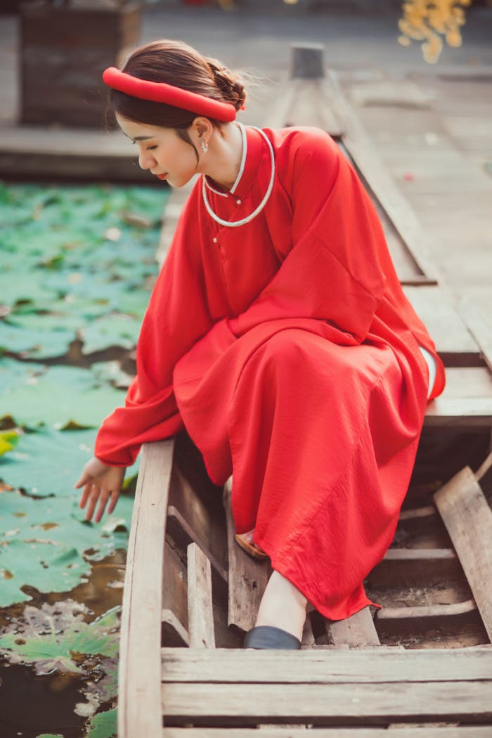 A woman in a vibrant red dress and headband sits gracefully on a wooden boat by a lotus pond.